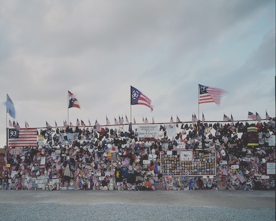 Chain link fence/gate that has been adorned with memorial material for Flight 93 and 9/11 generally. Small American flags line the top and larger flags are pinned up throughout alongside caps, firefighter jackets, clothing, photos, and other mementos.