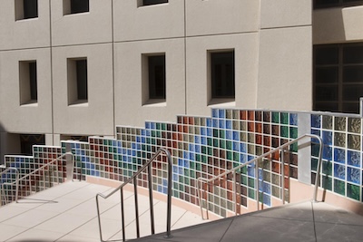 Outside photograph of a multi-colored glass wall descending alongside a cement stairwell. The glass wall takes up the bottom half of a building wall. The rest of the building&rsquo;s wall is a light tan color. 