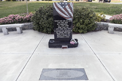 Black stone memorial resembling a large headstone that reads “If but one life be saved and one soul be comforted… all gave some some gave all and some still give. In honor and memory of all those who lost their lives in the rescue efforts of September, 11, 2001.” The stone is painted to look like an American flag is draped over the top.