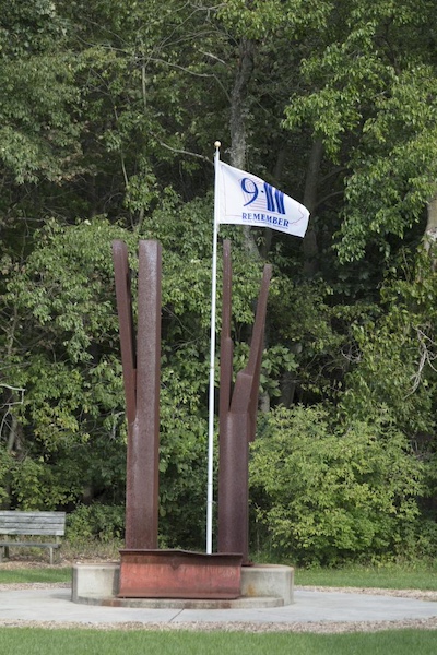 Two rusted steel beams like those from the Twin Tower rubble rise and each branch off to create four total branches. In the center, a white flag flies that reads “9/11 Remember New York City Washington DC Pennsylvania” with the outline of the Pentagon in the background