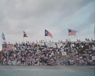 Chain link fence/gate that has been adorned with memorial material for Flight 93 and 9/11 generally. Small American flags line the top and larger flags are pinned up throughout alongside caps, firefighter jackets, clothing, photos, and other mementos. 