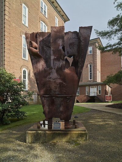 Sculpture of two hands holding another item in a green field with brick buildings in the background