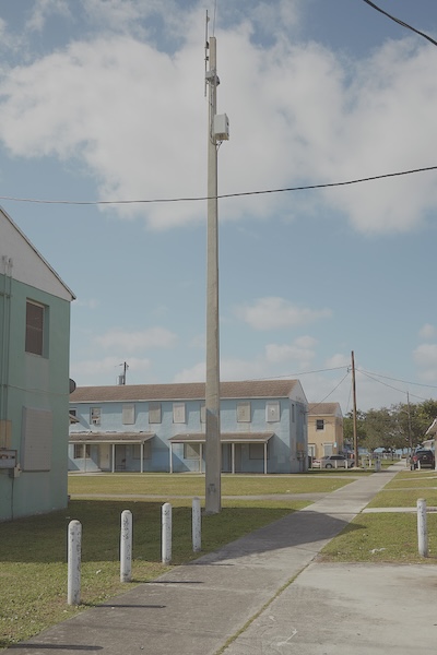 One full housing unit is in view, which is light blue, two stories, and has twelve windows and two doors. Two other housing units are partially visible, and a paved concrete path leads to the units. A telephone pole stands in front of the unit.