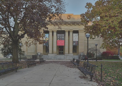 A wide walkway extends towards the museum, lined with black metal park benches and green space with trees behind them. Stairs ascend to the building entrance which is surrounded with columns. Between the columns, advertising posters are hung. 