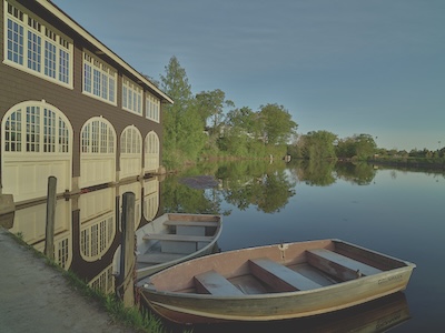 Boathouse on the lefthand side, and two small wooden boats float in the water next to it. Trees line the body of water into the distance of the photo.