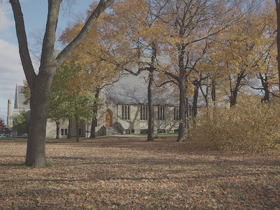 Side view of the church through a wooded area with tall trees and orang autumn leaves covering the ground.