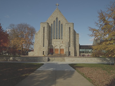 Front view of the chapel showing the cross at the top, stained glass windows ascending up, and three sets of orange double doors.