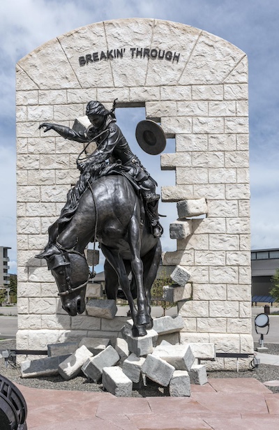 Statue of a person riding a horse through a white brick wall with the words "Breakin Through" on the top most part of the wall that is still intact.