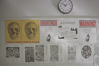 Bulletin board under an analog clock with various posters, pamphlets, and photographs pinned to it.