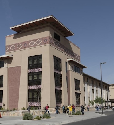 Outside of a tall, sand colored, four story building. Pedestrians walk by in the foreground.