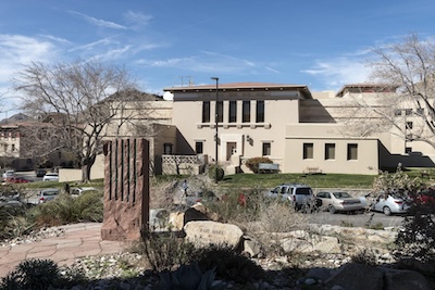 Desert garden sits in front of a large, light tan building with a flat roof. Cars are parked in a lot between the building and the desert garden.