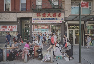 There are street vendors, resting pedestrians, and people walking by in front of a Chinese pharmacy.