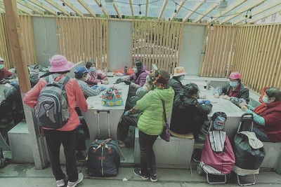 The women sit at tables with each other. The venue appears to be sunny and outdoors. Many of them have suitcases and backpacks with them.