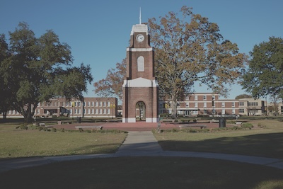 Large brick clocktower sits in a field of short green grass. Trees line the back of the clocktower, and more brick buildings sit behind the trees. There is a paved sidewalk leading to the clocktower.