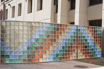 Outside image of a glass wall. The glass panes are colored to form a pointed arch. Outside to inside of the arch, the colors are blue, green, brown, orange, transparent, and then repeat. 