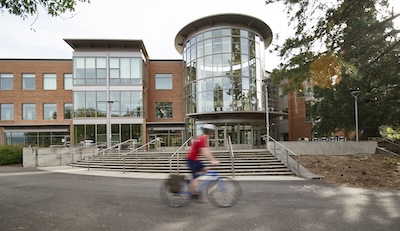 Front of a large brick and glass building. Steps lead up to the building, which has both a rectangular base and a cylindrical component that is completely glass. A man on a bicycle rides in front of the building. 