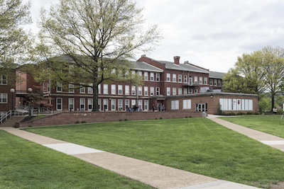 Dark brick three story building with rows of windows and a chimney just beyond a green lawn and some tall trees.