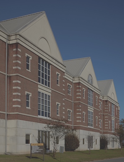 Large brick and cement building with three prominent gables. Windows are gridded on the front of the building. A small sign sits out front too far to read. 