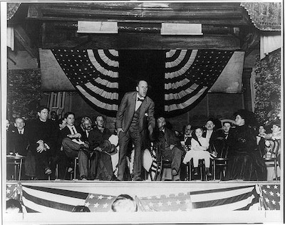 Eugene V. Debs making a speech on stage with two American flags and a group of people seated behind him
