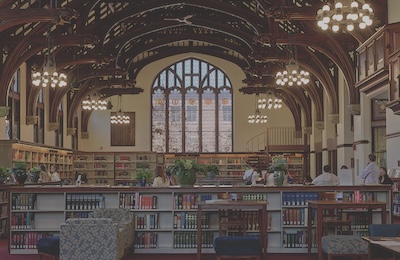 Inside of a library. Book shelves line the front and back walls, chandeliers hang from the ceiling, and there is a large window in the back of the room looking out onto a brick building. There is a small group of people in the library.