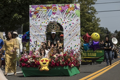 The float has lots of roses and flowers around the bottom and a crown floating at the top above a group of young girls who sit on the float and wave.