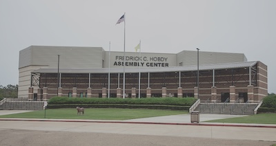 Outside of a large building that reads "Frederic C. Hobdy Assembly Center." The building contains two flags, the US flag and a smaller yellow flag. 