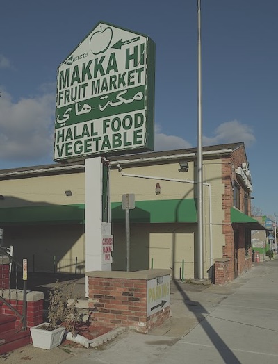 Sign in front of a building reads Makka Hi Fruit Market then Arabic script below that Halal Food Vegetable.