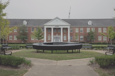 Outdoor setting with a brick building lining the back of the photograph. In the foreground, a large, horizontal sculpture of the letter G sits on a small patch of grass. The patch of grass is surrounded by a concrete path and a few benches. 