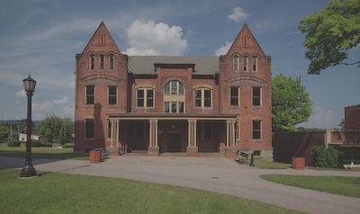 A large brick building sits at the end of a paved path. The building contains two gable roofs on each side of the building, and windows line the front. The sky is blue, with a few light white clouds