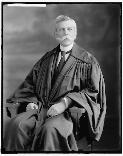 Portrait of Oliver W. Holmes in his judge robes. He has a large white handlebar mustache and is sitting in a chair.