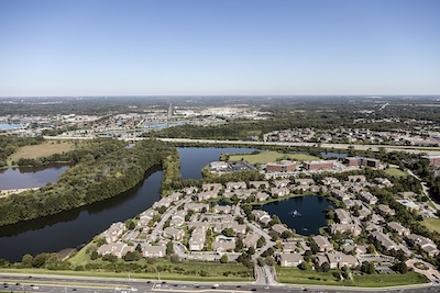 Outside aerial view of a private lake and the housing development that surrounds it. The sky is clear, and an interstate sits in front of the private community.