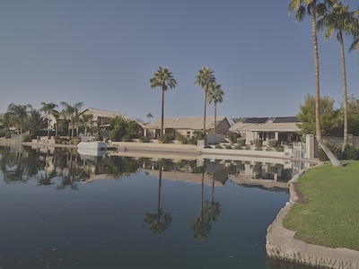 A large pond takes up most of the photo, and it is lined with palm trees, which are reflected in the pond. Toward the back of the photo are houses overlooking the pond.