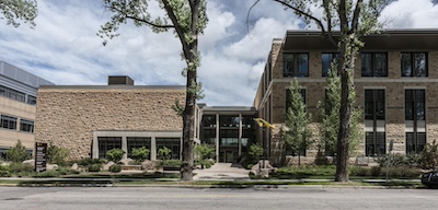 Shorter building on left connected to taller building on right by a glass hallway with entrance doors. Two tall trees and other green shrubbery grow in front.