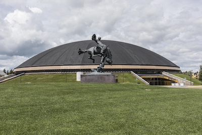 Weathered statue stands before a dome shaped building. The statue depicts a cowboy riding a bucking horse.