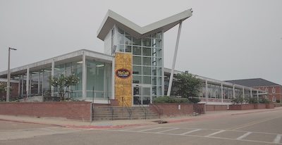 Outside of a one-story building with windows making up most walls. A window-walled tower rises above the rest of the building. The building sits off a paved road. 