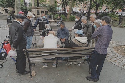 Elderly men gather around an outdoor table in New York City. Four of them sit across from each other on either side of the table on park benches and the rest of the men stand and observe.