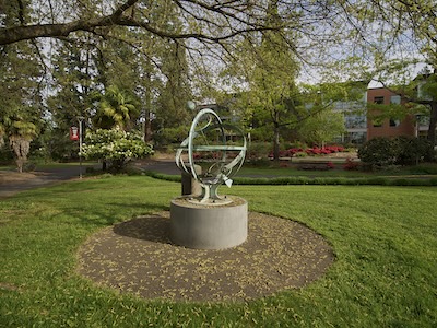 Outside garden area with grass and a few trees. In the center of the photo is a sculpture on a pedestal. The sculpture is orb-like, but transparent.