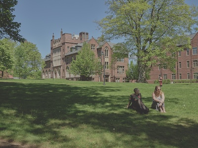 Two women sit on a grassy field with a few trees lining the perimeter. A large brick building sits in the background.