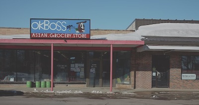 A small building with a OK Boss Asian Grocery Store sign. The sign also has a cartoon of a cowboy feeding a horse some green leaves.