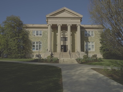 Outside front view of the library building. A concrete path cuts through a field leading up to the building. At the end of the path is a stairwell leading to the building. The building contains a gabled roof and four columns. 