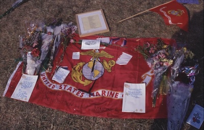 Red flag with notes, flowers, and photographs resting on top. Flag reads United States Marine Corps