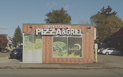 A small building has a sign reading Al-Qamar pizza and grill with pictures of pizzas on the windows