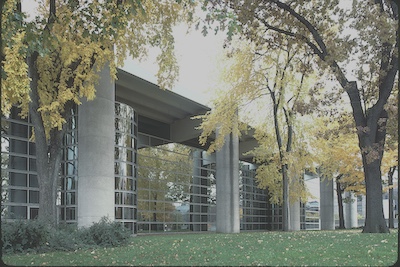 building with large cylindrical cement columns along the side to support it, the whole outside facing wall is reflective panes of glass. There are large trees with yellow leaves in the foreground