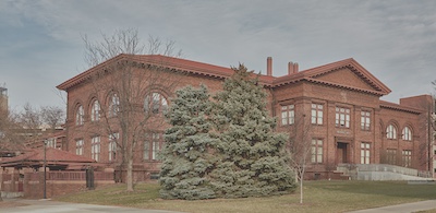 Orange or red brick building with a red roof. Picnic table with a red umbrella stands to the left and two large evergreen trees stand in front of the building.