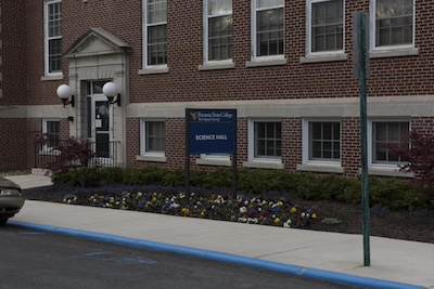 Brick building with rows of windows with a sign in front that reads "Potomac State College West Virginia University Science Hall". The doorway is framed by a decorative cement design and two large white ball sconces. Between the building and the sidewalk there are shrugs and a flower bed.