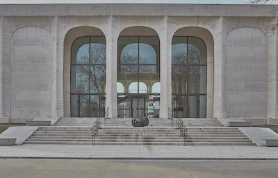 Steps with hand rails ascend to the entrance of the Sheldon Art Museum. There are three archways that stand in front of the large windows. On the steps there is a dark metal sculpture of a head on its side.