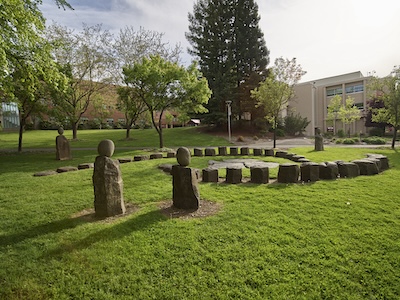 Stone sculptures sit in a grassy field with a few small trees. A brick building sits off the field in the distance. 