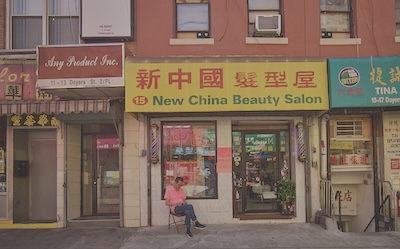 A man sits cross-legged on a chair in front of a store called New China Beauty Salon. 