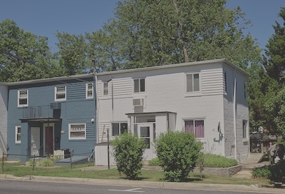 Outdoor view of two houses side by side. The house on the left is blue, and the house on the right is white. Both houses have flat roofs, two stories, and five windows. Grass and trees border the houses. 