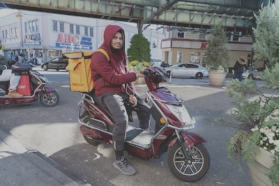 A man in a hoodie sits on a parked scooter with a large insulated backpack on his back